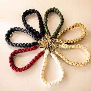 May include: A collection of seven paracord wrist lanyards arranged in a circular flower pattern. Each lanyard features a braided cord in colors including black, dark gray, olive green, tan, cream, and red, all attached to a brass-toned metal ring.
