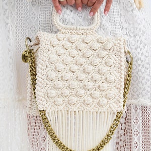 Puede incluir: Un bolso blanco de crochet con una correa de cadena dorada y detalles de flecos. El bolso tiene un diseño texturizado único con pequeños círculos en relieve.