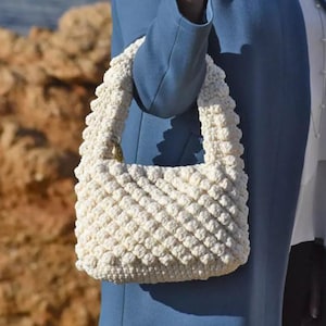 May include: A white crocheted handbag with a textured, puffy design. The bag has a short strap and a small, gold-colored clasp.