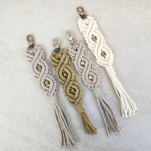 Op de afbeelding: Vier macramé sleutelhangers met kwastjes. De sleutelhangers zijn gemaakt van beige en gouden draad en hebben kleine gouden kralen.