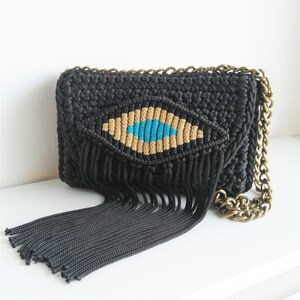 May include: Black crocheted crossbody bag with a fringe detail and a blue and gold eye design.