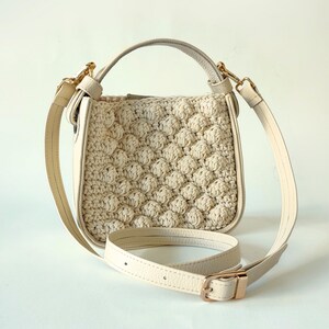 Op de afbeelding: Een crèmekleurige gehaakte handtas met een bovenste handvat en een lange verstelbare band. De tas heeft een textuur, geweven ontwerp.