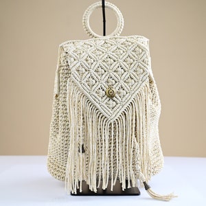 May include: A white crocheted backpack with a fringe design and a circular metal handle. The backpack has a button closure.