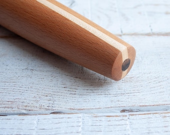 May include: A close-up of a wooden rolling pin with a light brown color and a white stripe running down the length of the pin. The rolling pin is on a white wooden surface.