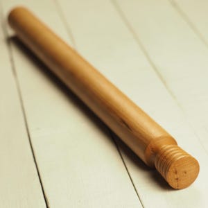 Könnte beinhalten: Ein hellbraunes Nudelholz aus Holz mit zylindrischem Körper und einem Gewindeende. Das Nudelholz liegt diagonal auf einer weißen Holzoberfläche. Die Holzmaserung ist sichtbar.