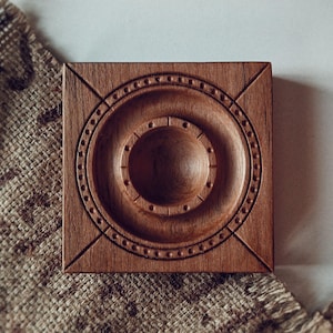 Könnte beinhalten: Quadratischer Holzuntersetzer mit einem geschnitzten kreisförmigen Design. Der Untersetzer besteht aus dunkelbraunem Holz und weist konzentrische Kreise und eine zentrale Vertiefung auf. Das Design umfasst kleine, gleichmäßig verteilte Punkte.