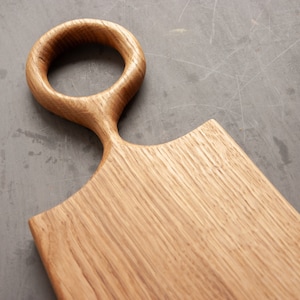 May include: A wooden cutting board with a unique, rounded handle. The board is made of light brown wood and has a smooth, polished surface.