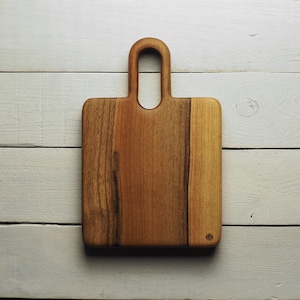 Può includere: Un tagliere rettangolare in legno con manico. La tavola è realizzata in legno marrone chiaro e scuro, con una superficie liscia e un manico arrotondato. Il tagliere è su una superficie di legno bianco.