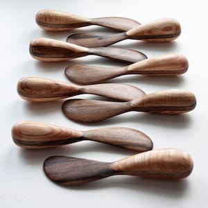 May include: A set of eight wooden butter knives with a natural wood finish. The knives have a unique design with a curved handle and a rounded blade.