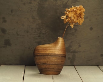 Peut inclure: Un vase en bois artisanal avec un design incurvé unique. Le vase est fait de bois en couches, mettant en valeur les motifs naturels du grain. Un arrangement de fleurs beiges séchées est placé dans le vase, ajoutant une touche de nature à la décoration.