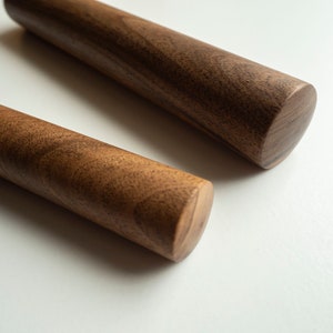 Può includere: Due mattarelli in legno con estremità arrotondate. I mattarelli sono realizzati in legno marrone scuro con motivi di venature visibili. I mattarelli sono di forma cilindrica e sembrano nuovi.