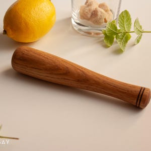 May include: A wooden cocktail muddler with a dark brown finish and two black rings around the end. The muddler is on a white surface.