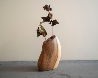 May include: A handcrafted wooden vase with a unique, curved shape. The vase is made of two tones of wood and holds dried flowers with brown leaves and small seed pods. The vase is displayed on a gray surface.