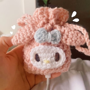 以下が含まれることがあります： 白い顔と青いリボンが付いたピンクの毛糸で編まれたキャラクターポーチ。ポーチには白い紐が付いています。