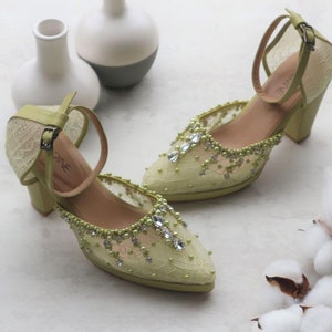 May include: A pair of light green high-heeled shoes with a delicate lace design and embellished with rhinestones and pearls. The shoes have a closed toe and an ankle strap.