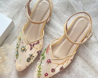 May include: A pair of beige slingback heels featuring a pointed toe and a sheer mesh upper adorned with intricate floral embroidery in shades of green, red, and gold, accented with small beads and pearls. The brand name Yevs is printed on the insole.