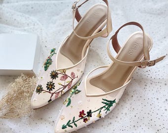 May include: A pair of cream-colored pointed-toe slingback heels featuring sheer mesh panels adorned with intricate floral embroidery in shades of red, green, and pink, accented with small beads. The insoles are stamped with the brand name Yevs.