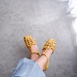 K&ouml;nnte beinhalten: Ein Paar gelbe geflochtene Sandalen mit Kn&ouml;chelriemen. Die Sandalen werden von einer Person getragen, die auf einem Betonboden sitzt.