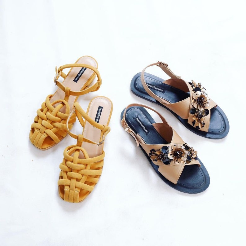 K&ouml;nnte beinhalten: Zwei Paar Sandalen. Ein Paar ist gelb mit einem gewebten Design. Das andere Paar ist braun mit einer schwarzen Sohle und mit goldenen Blumen verziert.