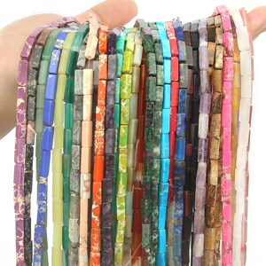 May include: A selection of rectangular gemstone beads in a range of colours, such as purple, green, orange, and blue. The beads are strung together, displaying diverse textures and patterns. The beads are held up against a white backdrop.