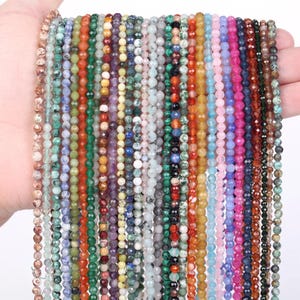 Könnte beinhalten: Eine Sammlung mehrerer vertikaler Stränge aus kleinen, facettierten, runden Edelsteinperlen in einer Vielzahl von Farben, darunter Rot, Orange, Gelb, Grün, Blau, Violett, Rosa, Braun, Weiß und Schwarz, die parallel nebeneinander angeordnet sind.