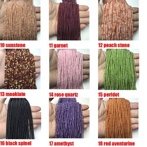 May include: Assortment of 18 strands of gemstone beads, each strand is a different color and type of gemstone. The beads are small and round. The strands are laid out in rows of three, with each row labeled with a number and the name of the gemstone. The gemstones include sunstone, garnet, peach stone, mookiate, rose quartz, peridot, black spinel, amethyst, and red aventurine.