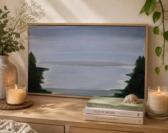 May include: A framed landscape painting with a serene lake scene in shades of blue and gray. The artwork is displayed on a wooden surface, alongside lit candles, books, and decorative elements, creating a cozy, inviting atmosphere.