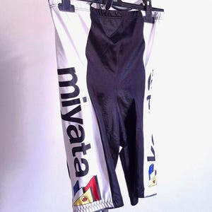 以下が含まれることがあります： 「Miyata」のブランド名がプリントされた、白と黒のサイクリングショーツ。ショーツは黒いパネルとカラフルなアクセントが特徴です。クロム製のハンガーに吊るされています。この衣服はおそらく伸縮性のある素材で作られています。
