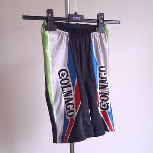 May include: Black and white cycling shorts with the word "COLNAGO" printed on the sides. The shorts have blue, red, and green accents. The shorts are hanging on a hanger.