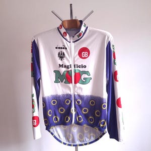May include: White cycling jersey with a full zipper, featuring the text "Maglificio M G" in green and red. The jersey has blue and purple accents, with red circles and the letters "GB".