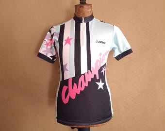 May include: A short-sleeve cycling jersey with a half-zip collar, featuring a light blue, black, and white vertical striped pattern on the upper chest, pink stars, and the word "Champion" in large pink script across the black lower section.