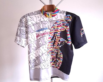 May include: A short-sleeved cycling jersey with a colorful design. The left side is white with gray text, while the right side is black with a cyclist graphic. The collar has a rainbow stripe design and a zipper.