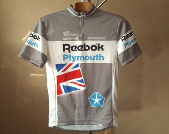 Może przedstawiać: Szary i biały jersey kolarski z napisem "Reebok Plymouth" i wzorem flagi Unii Jack. Jersey ma zamek błyskawiczny i jest wykonany przez Shimano.