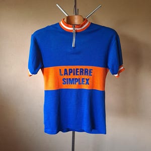 May include: A blue and orange vintage cycling jersey with the text "LAPIERRE SIMPLEX" across the chest. The jersey has a white and orange striped collar and sleeve cuffs, and a partial zipper.