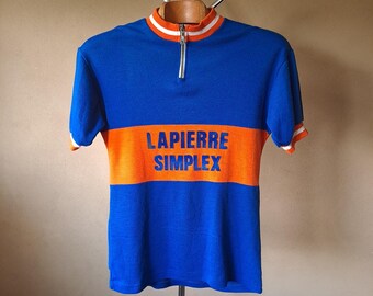 May include: A blue and orange vintage cycling jersey with the text "LAPIERRE SIMPLEX" across the chest. The jersey has a white and orange striped collar and sleeve cuffs, and a partial zipper.