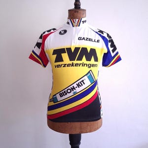 May include: A vintage cycling jersey in yellow, white, black, red, and blue, displayed on a mannequin. The jersey features the text "TVM verzekeringen" and "GAZELLE" with a "BISON-KIT" logo. The jersey has a short zipper and short sleeves.