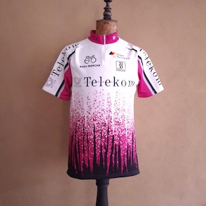 Pode incluir: Uma camisola de ciclismo de manga curta branca e magenta com os textos Telekom, Team Telekom e Eddy Merckx. A peça tem uma gola com meio fecho, detalhes pretos na bainha e um padrão gráfico de riscas verticais magenta e pretas.