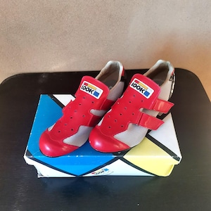May include: A pair of red cycling shoes with white mesh panels and velcro straps. The shoes have the word "LOOK" printed on the side in a colorful logo. The shoes are sitting on top of a colorful box.