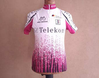 May include: A white and magenta short-sleeve cycling jersey featuring the Telekom logo, Eddy Merckx branding, and a Team Telekom emblem. The design includes a gradient pattern of magenta vertical streaks fading into white, a high collar, and a front zipper.