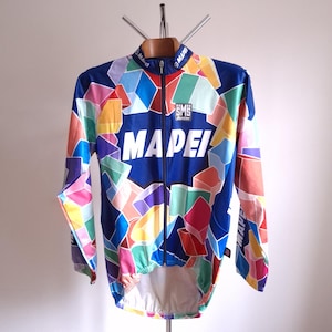 Op de afbeelding: Een wielershirt met lange mouwen en een levendig geometrisch patroon in blauw, roze, oranje, groen en paars. Het shirt heeft het woord "MAPE" in grote witte letters over de borst en hangt aan een houten kleerhanger.