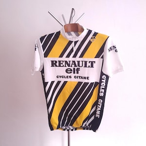 以下が含まれることがあります： 白、黄色、黒のヴィンテージサイクリングジャージ。"RENAULT elf CYCLES GITANE"のロゴ入り。ジャージは短いジッパーと半袖。デザインには斜めの黒いストライプと袖の"elf"ロゴが含まれています。