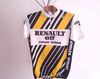 May include: A vintage cycling jersey in white, yellow, and black, featuring the "RENAULT elf CYCLES GITANE" logo. The jersey has a short zipper and short sleeves. The design includes diagonal black stripes and the "elf" logo on the sleeve.