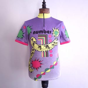 Può includere: Una maglia da ciclismo viola a maniche corte con colletto giallo e cerniera frontale. La grafica presenta la parola number, un grande numero uno e un design a pellicola gialla con sagome nere di ciclisti, stelle verdi e cerchi rosa.
