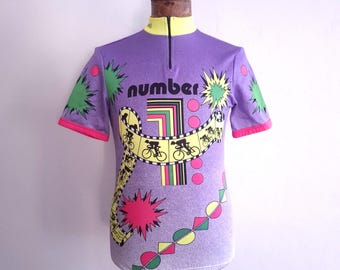 May include: A short-sleeve purple cycling jersey with a yellow collar and a front zipper. The graphic features the word number, a large numeral one, and a yellow film strip design with black silhouettes of cyclists, green starbursts, and pink circles.