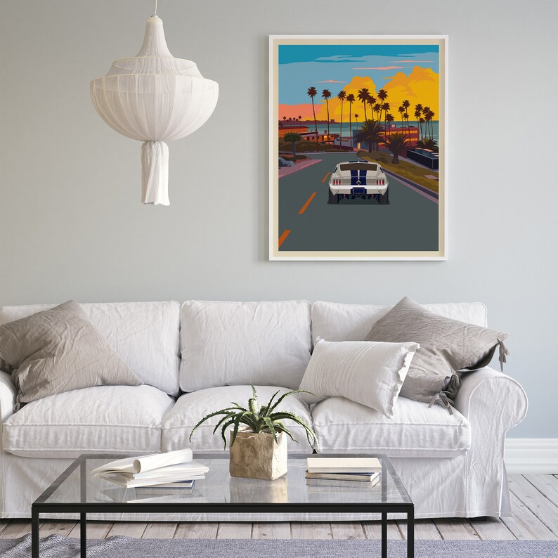 K&ouml;nnte beinhalten: Ein wei&szlig;es Sofa mit zwei Kissen und einer Pflanze auf einem Glas-Couchtisch vor einem gerahmten Druck eines wei&szlig;en Autos, das eine Stra&szlig;e mit Palmen und einem Sonnenuntergang im Hintergrund entlangf&auml;hrt.