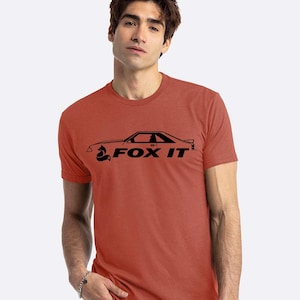 May include: A rust-colored t-shirt featuring a black graphic of a car and the text "FOX IT". The design is centered on the front of the short-sleeved shirt. The shirt is made of a soft material.