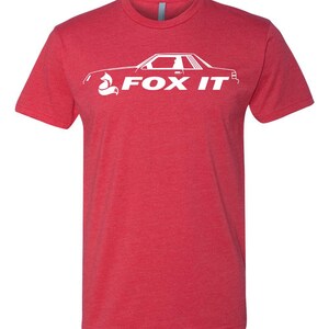 May include: Red t-shirt with a white graphic of a car and the text "FOX IT". The design features a stylized fox head incorporated into the car's silhouette. The shirt has short sleeves and a crew neck.