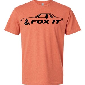 May include: A coral-colored t-shirt featuring a black graphic of a car silhouette with the text "FOX IT". The design includes a stylized fox head. The shirt has a crew neck and short sleeves.
