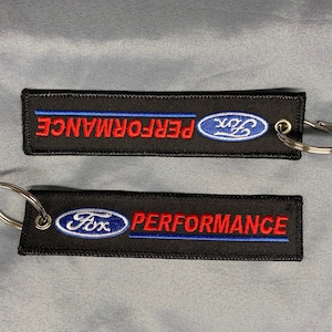 May include: Two black keychains with silver key rings. Both keychains have a blue oval with the word "Ford" in white text and the word "Performance" in red text. The top keychain has a blue stripe above the text.
