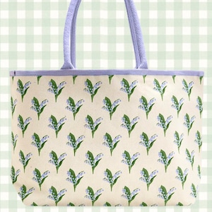 May include: A canvas tote bag with a floral pattern of blue and green lily of the valley flowers on a cream background. The bag has a lavender trim and matching handles.